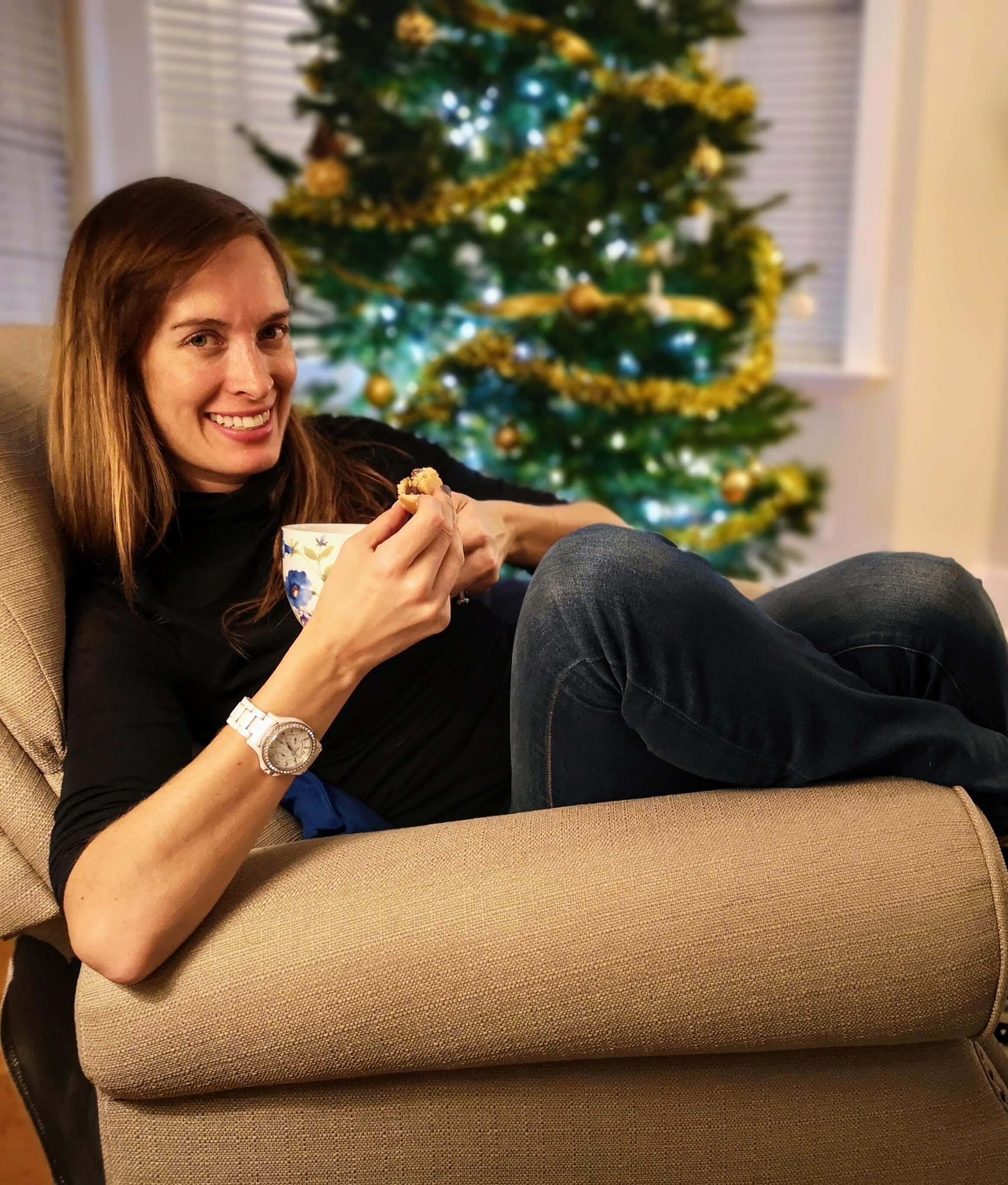 Breanna sitting in a chair with a hot drink and cookie, in front of a Christmas Tree.